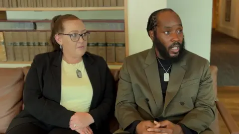 A woman wearing a yellow top and a black blazer and a man wearing a green blazer and a black top, sitting on a sofa and looking at something off-camera.