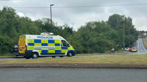 A police van is parked on a grass roundabout. Ahead are three cars including a police car in a crashed position on a road. 