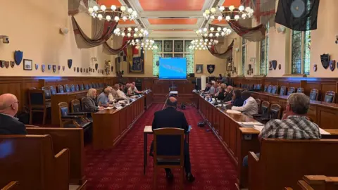 LDRS A wide shot of a the inside of Bootle Town Hall. The floor is a crimson red. There are long desks either side with people sat in chairs behind them facing each other. There is a man sitting in a chair in the middle of the floor looking out towards everyone else.
