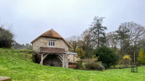 A stone mill is the focus of the picture with the wooden wheel visible underneath a tiled roof. There are other stone buildings behind it with gardens. One has a child's swing in the garden. The grass in front of the mill is a very vivid green colour while the sky is a very pale blue.