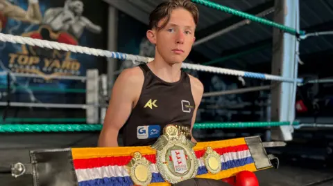Emma Buagh/BBC Jerry-Mike wearing red boxing gloves and sitting on the boxing ring with his championship belt.
