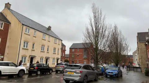 A new build estate with cream rendered and red brick townhouses where there are lots of cars parked on the road. It is raining, the road is wet and the sky is grey and cloudy.