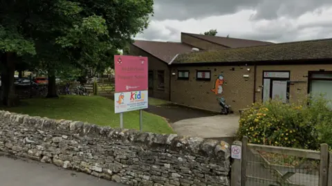 The entrance to Middleham Primary School. A dry-stone wall runs across the front of a low-rise brick building with a wooden gate and pathway leading into the school grounds. A large pink sign on two metal posts reads: Middleham Primary School. Below it is advertising for Kidz Day Nursery.