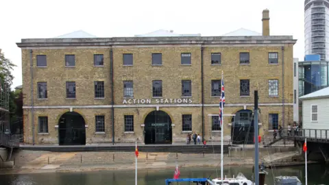 Google A naval sand-coloured building with arches and the words "action stations" above the front entrance. It is adjacent to a harbour with sailing boats.