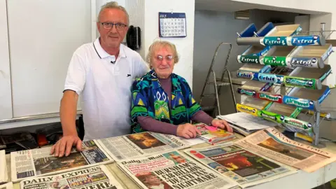 A man in glasses with blond hair, wearing a white polo shirt, stands behind a newsagent's counter next to a shorter woman in a patterned green, blue and yellow shirt over a purple long-sleeved top. She has blonde hair cut short and glasses on. To their right is a stand displaying chewing gum.
