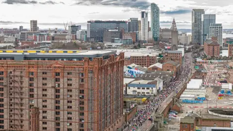 Stratus Imagery Limited A line of runners race down a road past a Victorian multi-storey brick dockhouse with the city centre and high-rise buildings ahead.