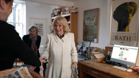 PA The Queen is shaking a woman's hand inside a book store. The book store has picture on the wall which says "the historic city of bath. Next to the Queen is a desk with a flat screen computer. Behind Her Majesty a woman is wearing a pink and green scarf is smiling. 