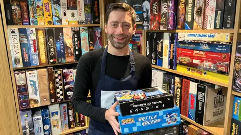 A man is standing in front of shelves containing hundreds of board games. He is wearing a black top with navy blue apron, and has dark brown hair with dark brown stubble. He is holding two board games in his hands and is smiling at the camera