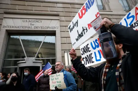 Reuters Protesters carrying signs outside of US Agency for International Development building One sign reads: Musk & Trump, Keep your fascist hands off USAid