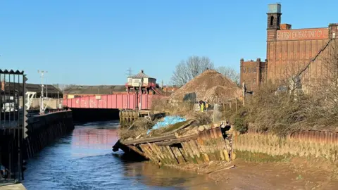 A shot of the River Hull showing the collapsed pilings on one bank of the river. There is a red bridge in the background