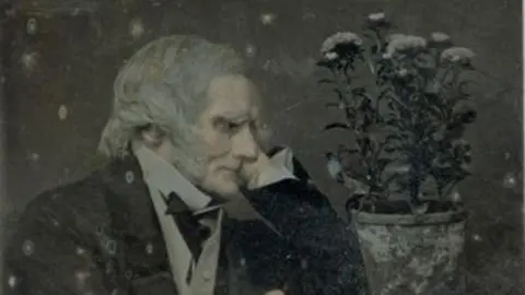 The image shows an elderly man with grey hair wearing a suit and looking at a pot of flowers.