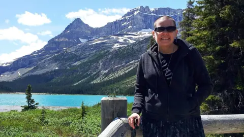 Cindy Baldassi is stood in front of a blue lake and towering mountain peaks with snow visible at highest points
