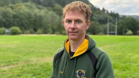 BBC Tudur Roberts stood on a rugby pitch with trees behind it. He has short ginger hair and is wearing a green and yellow hoodie.