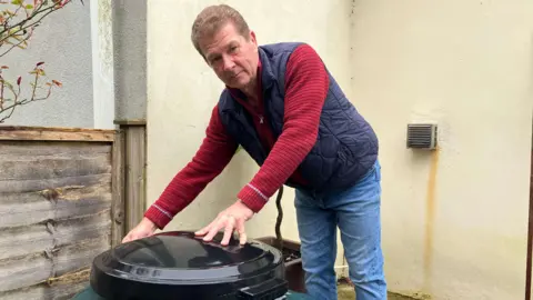 A middle-aged man with brown hair and a gilet stands over a heating oil tank