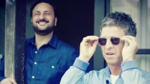 Nick Brine and Noel Gallagher. Noel is wearing sunglasses, a blue jacket and a gold chain bracelet. Nick has dark hair and a beard and is wearing a dark blue shirt. 