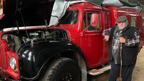 Edd Smith/BBC Richard Wallis wearing a checked shirt with a black fleece waistcoat with a flat cap on. He is standing next to the red and black rapid intervention vehicle, which has its bonnet partially open.