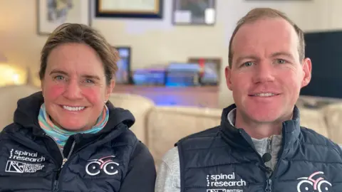 James Burridge/BBC Piggy and Tom March sitting side by side on a cream sofa, both wearing dark blue gilets with Spinal Research and British Eventing Support Trust logos on one side of the chest, and a cycling logo on the other. A table lamp, sideboard, framed pictures on the wall and a TV can be seen behind them. 