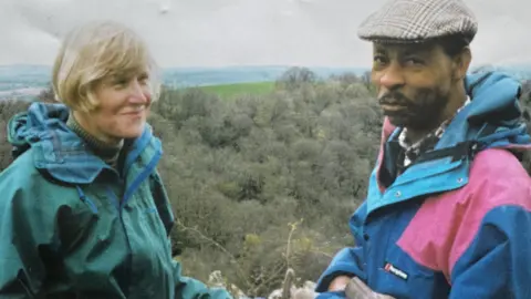 Lythan Stephenson The couple pose together in a photo taken during an outdoor walk. They are both wearing waterproof jackets and appear to be on top of a hill, or at height.
There are rolling hills and trees behind them.