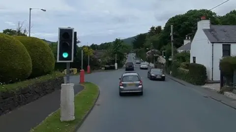 The traffic lights at the junction in Marown. The light is green and there are cars on the road, with houses on the right and green shaped hedges on the left.
