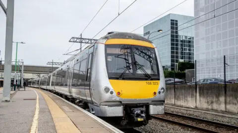 East West Rail A silver and yellow train at Milton Keynes railway station, showing track buildings to the right and power lines.