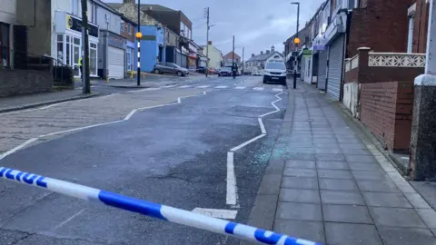 Julie Smith/BBC Police tape is stretching across a street with a police van and officers further up the street.