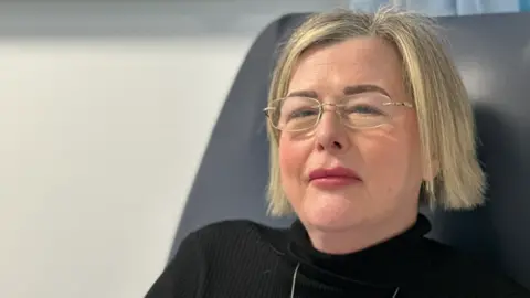 BBC A woman with blonde hair and glasses is looking at the camera. She is sitting down on the right of the photo and wearing a black top.
