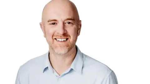 David Powles David Powles is wearing a light blue shirt and is smiling. He stands against a white background.