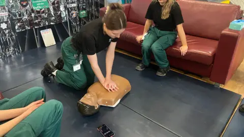 A girl with brown hair in a claw clip is demonstrating how to do CPR. She is wearing a black shirt and green trousers. She is pushing down on the chest of a CPR mannequin 