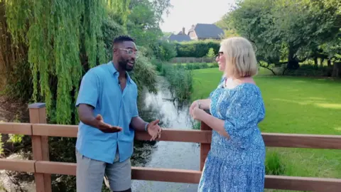 Tinie Tempah in a blue shirt, glasses and shorts on the left and Sophie Law in a blue dress and glasses on the right and they are chatting on a bridge over a river in a green area.