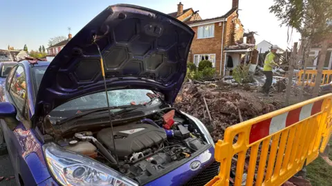 A blue car with it's hood up. The car is surrounded by rubble and a yellow cordon. The damaged houses can be seen in the background.