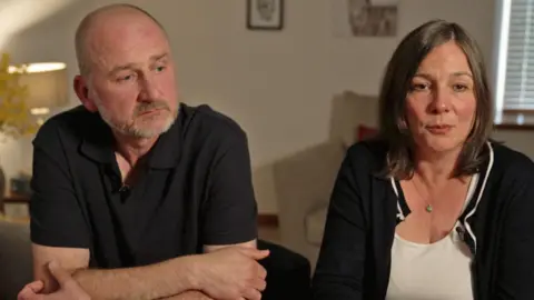 Mark Dowey in a black polo shirt leans on a table looking off to the side in thought, beside his wife Ros in a white top and black cardigan who is looking into the camera. They sit in a light lounge with a lamp on in the background and the blinds drawn.