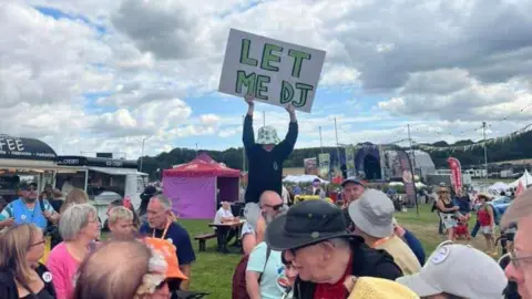 Family handout A young boy wearing a green and white bucket hat, a black sweatshirt and light grey shorts. He's sitting on the shoulders of a bald man and holding up a large white sign saying 'let me DJ' on in green letters. He's at a festival in the daytime, and there are crowds of people, tents and bunting all around him.