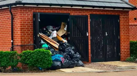 Johnny Luk A brick-built bin store beside a residential building is overflowing with rubbish, with black refuse sacks, wooden planks and other waste piled high and spilling out through an open door onto the pavement. A second bin store door remains closed.