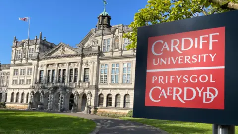 A red sign reading “Cardiff University / Prifysgol Caerdydd” stands prominently in the foreground. Behind it, a wide path curves toward a grand stone university building with a central tower and arched entrance. The surrounding lawn is green and sunlit, with trees framing the scene.