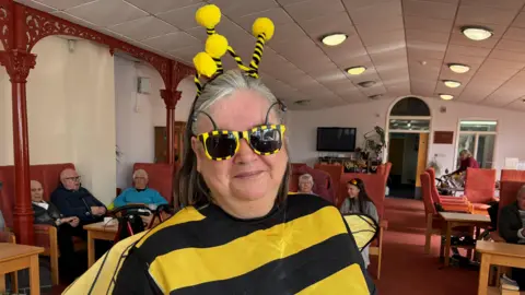 A woman in a fancy dress bee outfit and black and yellow sunglasses smiles at the camera , behind her are a group of elderly people sitting in chairs at a day centre 