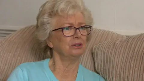 A woman with grey hair and glasses in a blue top, sat on a beige sofa