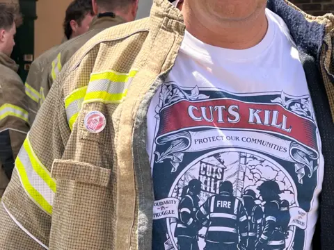 A man wearing a fire jacket and a white t-shirt. You cannot see the man's face. The t-shirt says "Cuts Kill, Protect Our Communities". There are three other men standing in the background.