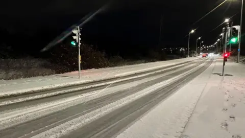 Snow and slush on the road. There is a set of traffic lights and cars in the distance. It is early morning and still dark.