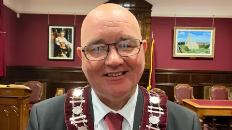 William Walker is smiling and wearing glasses. He is wearing a red collarette and a grey suit with a red tie.