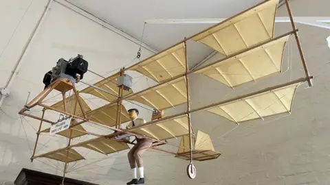 A replica model of the Pilcher triplane is suspended from a white wooden arm, attached to the wall.