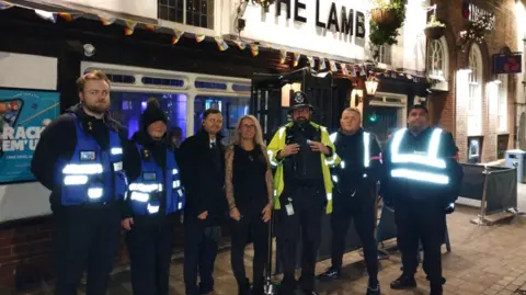 Seven people standing outside a pub at night, called the Lamb, one in hi viz jacket.