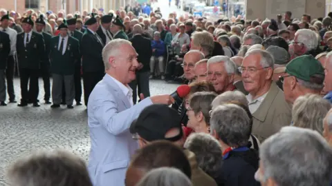 Stuart Woodward/BBC Dave Monk wears a light blue jacket and white shirt. He holds a microphone with a red cover and he is speaking to a crowd of people. Behind Dave is a group of men in uniform who are about to play the Last Post at the Menin Gate in Ypres, Belgium