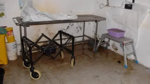 CPS The corner of a mortuary room at Elkin and Bell Funerals in Gosport. There is a metal trolley on wheels pushed up against a moldy white wall with a rumpled plastic sheet on it. Three tubs of chemicals are piled on the floor on the left hand side. The floor is grey and filthy with brown wet patches. 