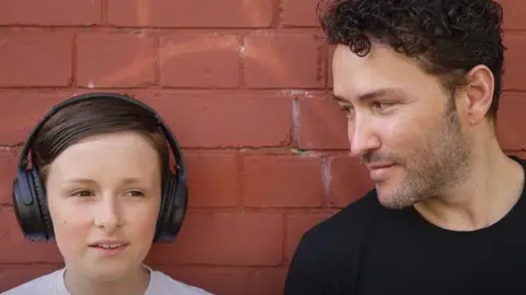 Richard and Jaco standing against a brick wall. Jaco is wearing headphone. 