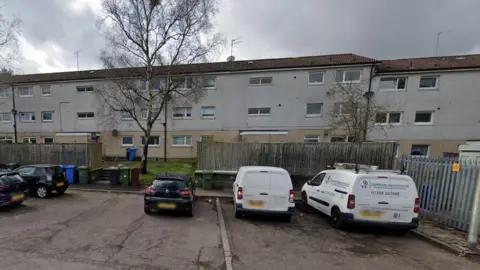 Cars are parked outside a block of grey three-storey flats. The ground floors are obscured by a tall wooden fence. 