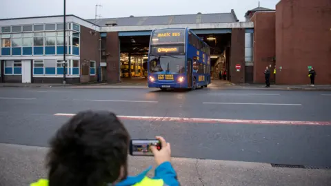 Stagecoach A man films a Magic Bus drawing out of a garage through his smartphone. 