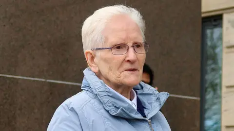 Spindrift Head and shoulders shot of Marie O'Gorman, who has short grey hair swept back, outside Glasgow Sheriff Court. She is wearing a light blue jacket with a large collar and has dark-rimmed glasses. She is looking ahead, to the right of the photographer.
