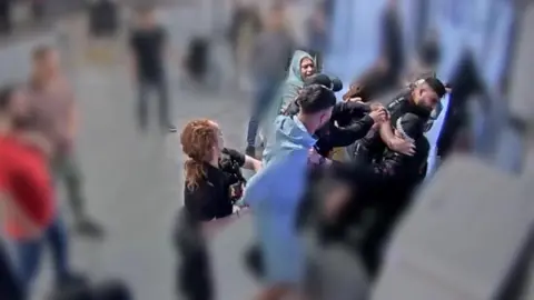 PA Media A still of a group of police officers and others mid-brawl close to pay station indoors. Large parts of the image are blurred to protect the identity of others. 