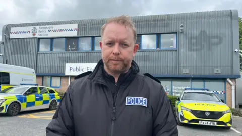 Andy Wills looking at the camera. He is wearing a black jacket and on the right there's a blue police badge. He is stood in front of the Barnstaple police building which has two parked vehicles.