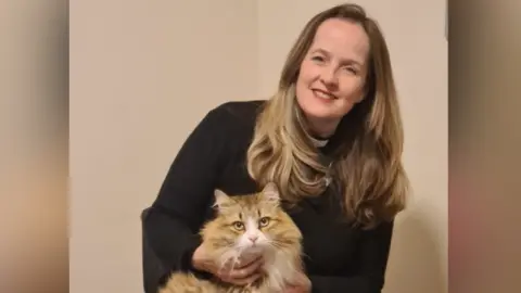 Jennifer Adams-Massmann The Reverend Jennifer Adams-Massmann is holding Rory, a golden and white cat, on her lap. She is wearing a black gown. She has long dark blonde hair and is sat in front of a wall.